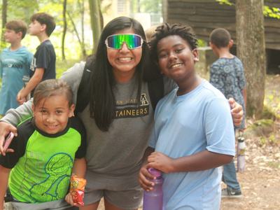 Three children with counselor outdoors; counselor's shirt reads "TAINEERS".