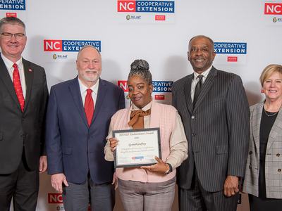 Five people standing; center woman holds a certificate; backdrop reads "NC Cooperative Extension"