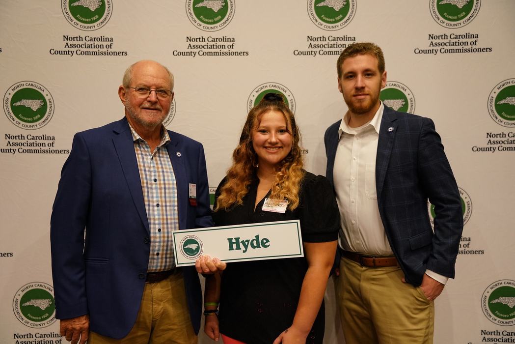 Three people standing before North Carolina County Commissioners backdrop holding sign "Hyde"