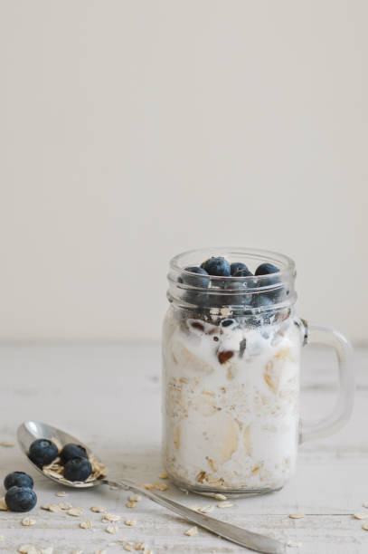 Vertical photo of overnight oats with coconut milk and blueberry served on wooden table with a metal spoon. Porridge in a jar. Breakfast meal
