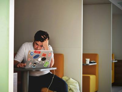 A man working on a laptop, holding his head in a gesture indicating anxiety and stress.