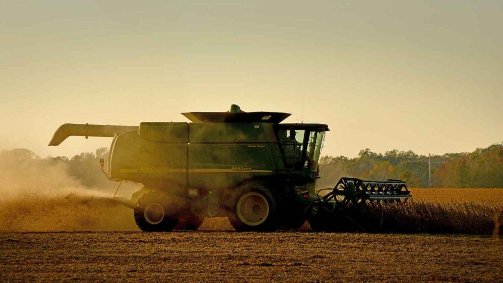 A green combine harvester in a field during sunset, creating dust clouds as it harvests crops.