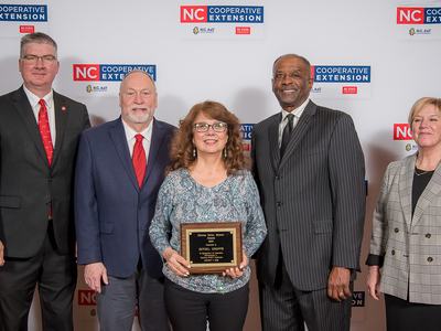 Five people at NC Cooperative Extension event; center woman holds an award plaque.