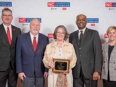 Five people standing at NC Cooperative Extension backdrop; center woman holds award plaque.
