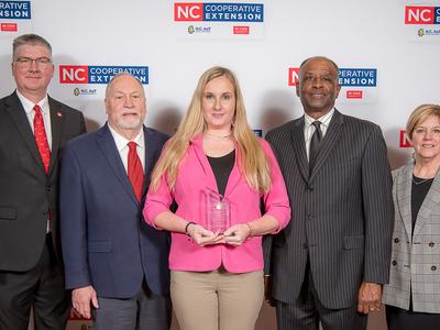 Woman holding glass award, four others standing before "NC Cooperative Extension" backdrop