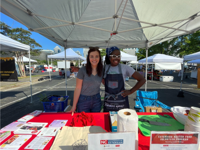 Two women at a farmers market booth under a canopy with "NC Cooperative Extension" sign.