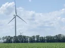 Wind turbines rising above a green crop field with a tree line and cloudy sky