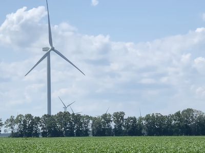 Wind turbines rising above a green crop field with a tree line and cloudy sky