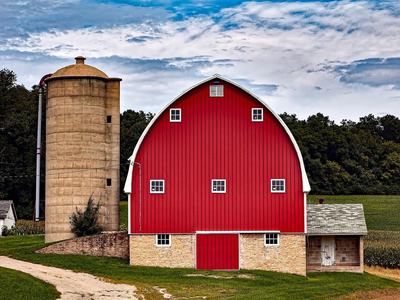Red gambrel barn with stone foundation beside tall concrete silo and dirt driveway