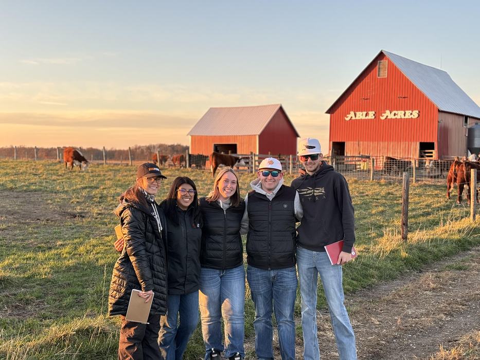 Five people posing in front of red barn labeled "ABLE ACRES" on a farm at sunset