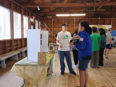 Teen in white 4-H shirt speaking with person in blue jacket at indoor display table