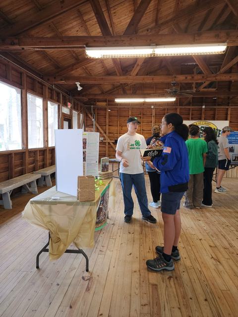 Teen in white 4-H shirt speaking with person in blue jacket at indoor display table