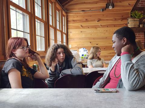 Four teenagers seated at a table in a wood-paneled room talking, "Pure Leaf" bottle visible.