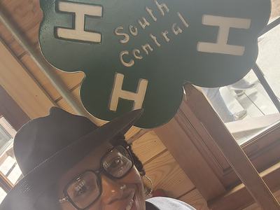 Person wearing hat and glasses beside green four-leaf sign reading South Central