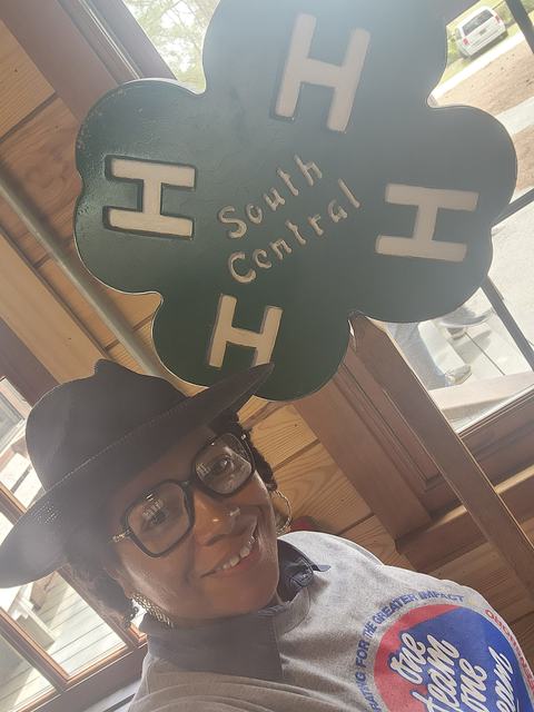 Person wearing hat and glasses beside green four-leaf sign reading South Central