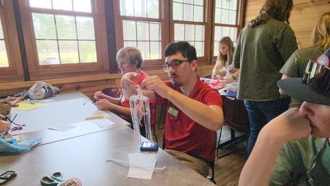 Young man in red shirt crafting a yarn tassel with hanging strands at a table