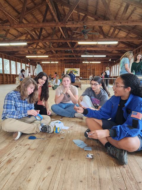 Group of five teens sitting on a cabin floor making crafts with tape, paper, and scissors