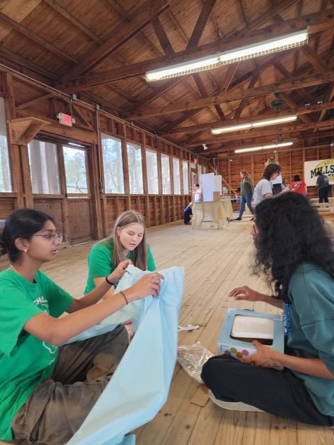 Three girls sitting on a wooden floor assembling light-blue fabric and a small box