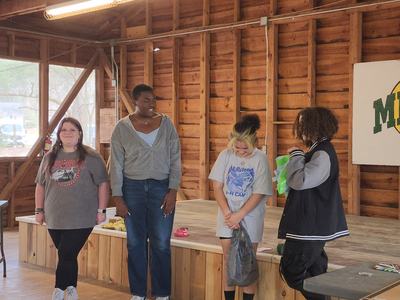 Four teens standing on a wooden stage inside a cabin, sign reading "MILLSTONE".