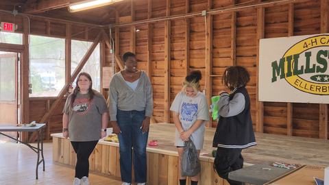 Four teens standing on a wooden stage inside a cabin, sign reading "MILLSTONE".