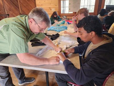 Adult guiding a young person working on a white T-shirt at a long table in a wooden room