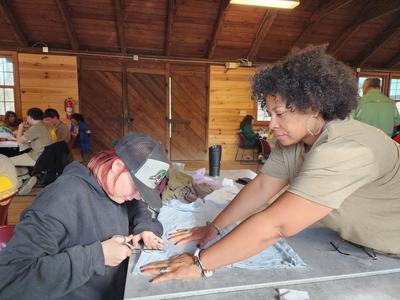 Person cutting fabric with scissors while another person holds it on a table in a wooden cabin