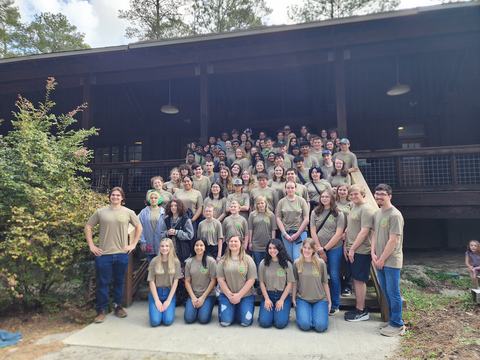 Large group of people in matching tan T-shirts posing on cabin steps outdoors