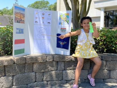 A young girl sits on a stone retaining wall beside her 4-H presentation trifold board at the Cloverbud Presentation Competition. She is smiling, holding a stick to point at her board, and displaying a green ribbon in her left hand, indicating her achievem