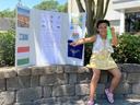 A young girl sits on a stone retaining wall beside her 4-H presentation trifold board at the Cloverbud Presentation Competition. She is smiling, holding a stick to point at her board, and displaying a green ribbon in her left hand, indicating her achievem
