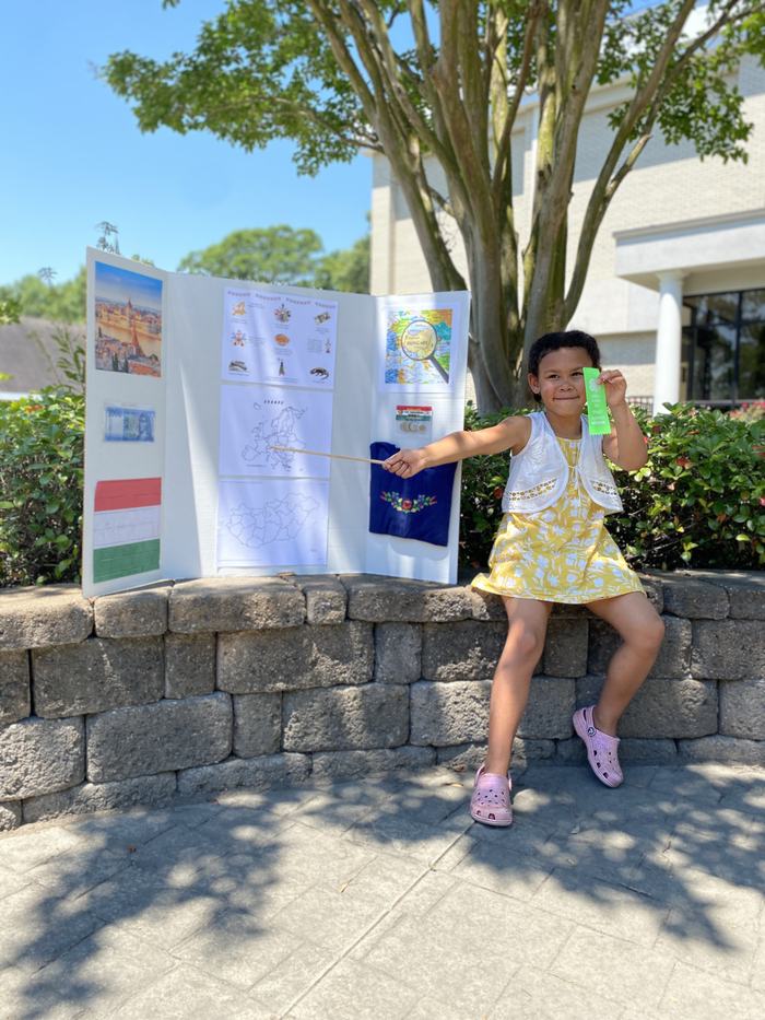 A young girl sits on a stone retaining wall beside her 4-H presentation trifold board at the Cloverbud Presentation Competition. She is smiling, holding a stick to point at her board, and displaying a green ribbon in her left hand, indicating her achievement in the competition.