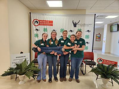 Five teens in green NC State 4‑H shirts; four hold a teammate horizontally under NC State 4‑H banner.