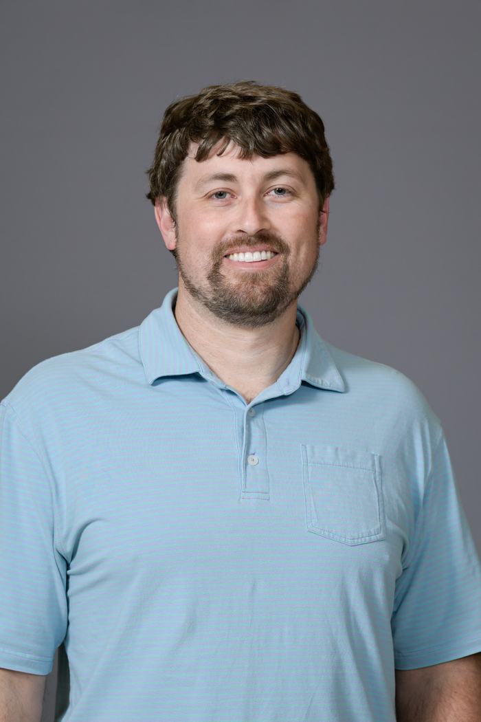 Adult man with beard smiling, wearing light blue polo against plain gray background