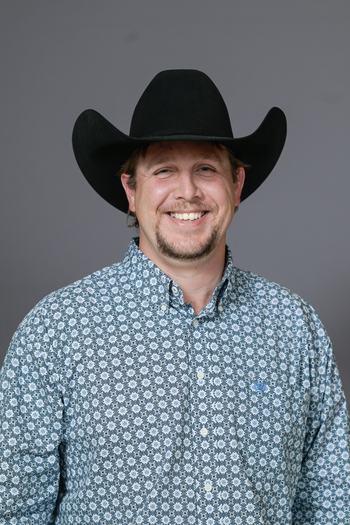 Man wearing black cowboy hat and patterned button-up shirt, smiling against gray background