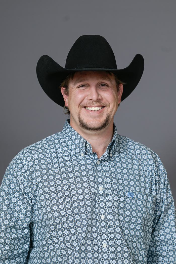 Man wearing black cowboy hat and patterned button-up shirt, smiling against gray background