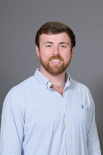 Young man with brown hair and beard wearing blue check button-down shirt against gray background