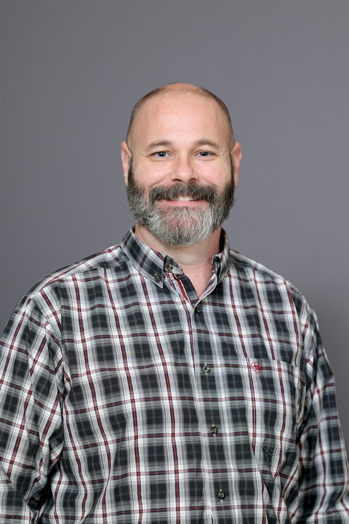 Bald man with beard wearing a plaid button-up shirt, facing camera against gray background