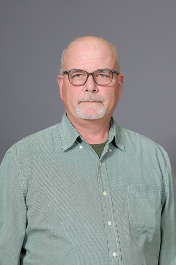 Head-and-shoulders portrait of a bald man wearing glasses and a green checkered button-up shirt.