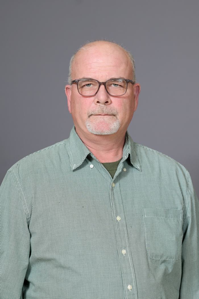 Head-and-shoulders portrait of a bald man wearing glasses and a green checkered button-up shirt.