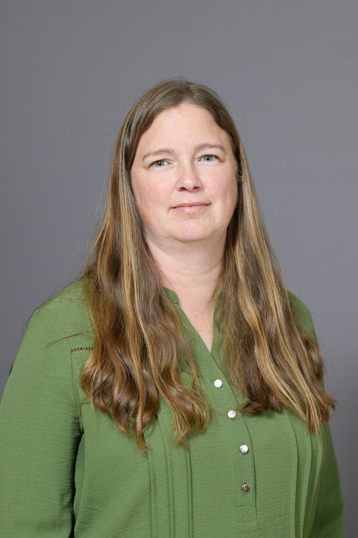 Woman with long brown hair wearing a green buttoned blouse, neutral gray background