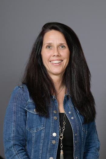Woman with long dark hair wearing a denim jacket and necklace, smiling at camera