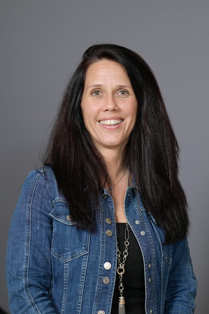 Woman with long dark hair wearing a denim jacket and necklace, smiling at camera