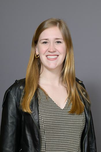 Woman smiling, wearing black leather jacket, houndstooth v-neck dress and gold hoop earrings
