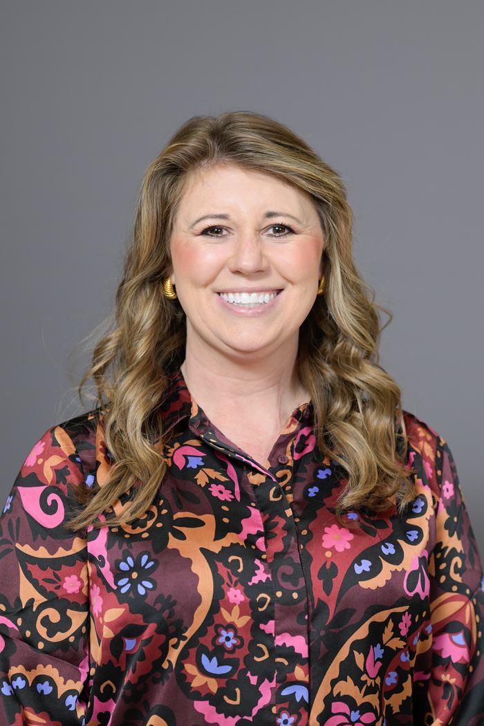 Woman with wavy blonde hair in patterned blouse, facing camera on gray background.