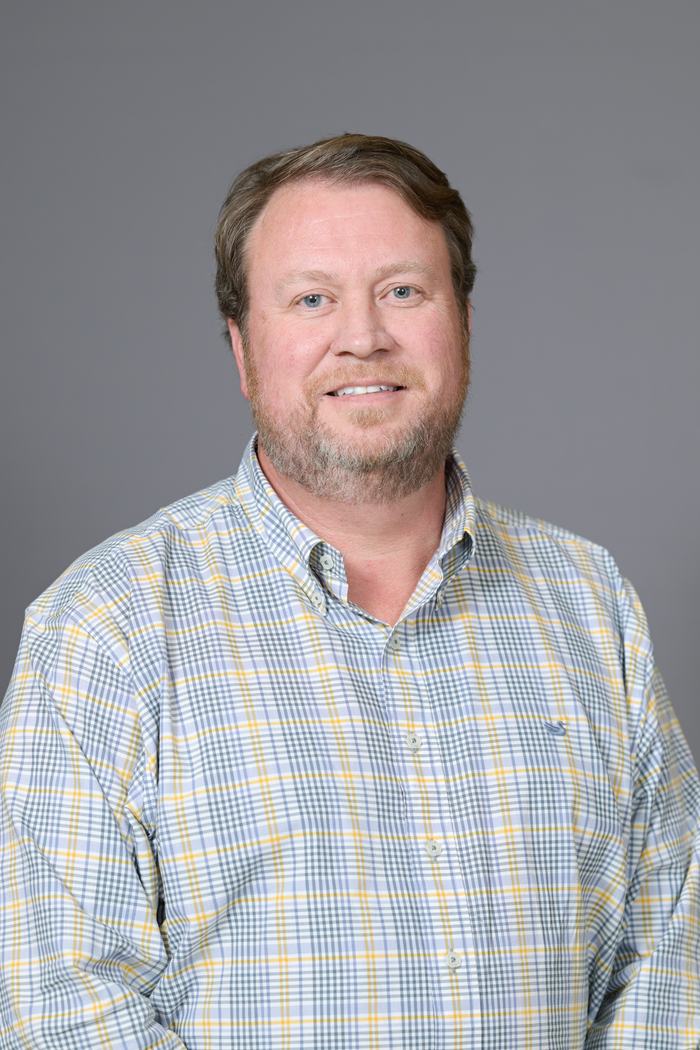 Adult man head-and-shoulders portrait wearing a plaid button-down shirt against gray background