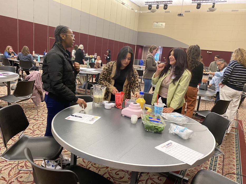 Three women at a round table preparing drinks with a blender and ingredients in a conference room