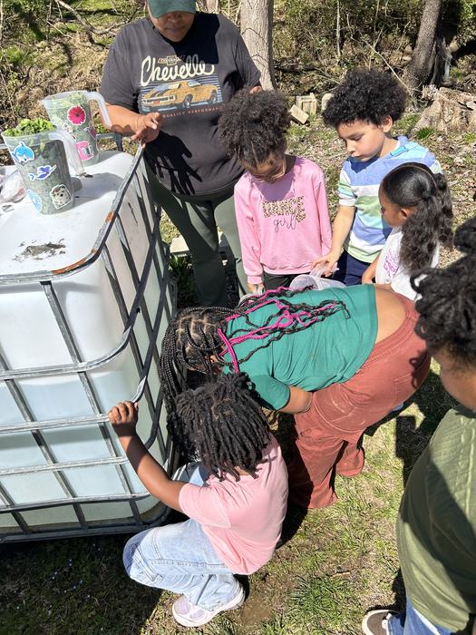 Adult and children around large water tank, child filling container; shirt reads Chevelle