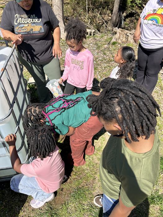 Children gathered around a large plastic water tank, one child holding a decorated plastic pitcher.
