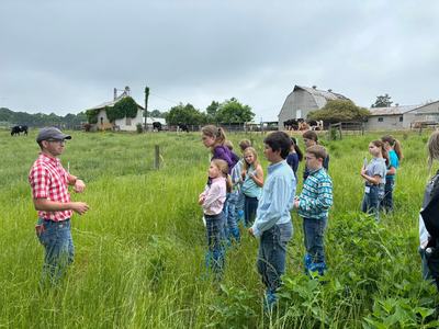 Man speaking to group of children standing in a grassy field near barns and grazing cows