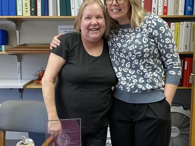 Two women standing side-by-side with arm around shoulder in an office; awards on table.