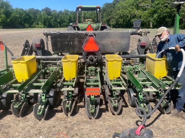 A farmer vacuuming out the corn planter attached to a tractor. The equipment features four yellow buckets for corn seed and eight bladed rows for soil cutting. The image is taken from the rear of the rig, showing both the corn planter and the back of the tractor.